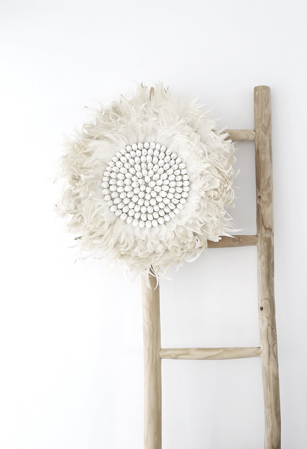 A handcrafted white feather juju wall decor with a beaded centre displayed on a rustic wooden ladder against a minimalist white background. A statement piece with soft textures and organic elegance
