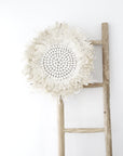 A handcrafted white feather juju wall decor with a beaded centre displayed on a rustic wooden ladder against a minimalist white background. A statement piece with soft textures and organic elegance