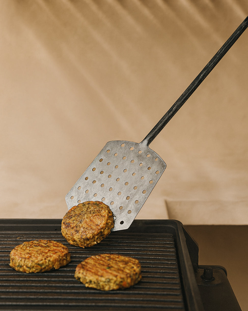 Forged stainless steel spatula flipping veggie patties on a grill, showcasing its textured handle and durable design in an outdoor barbecue setting