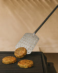 Forged stainless steel spatula flipping veggie patties on a grill, showcasing its textured handle and durable design in an outdoor barbecue setting