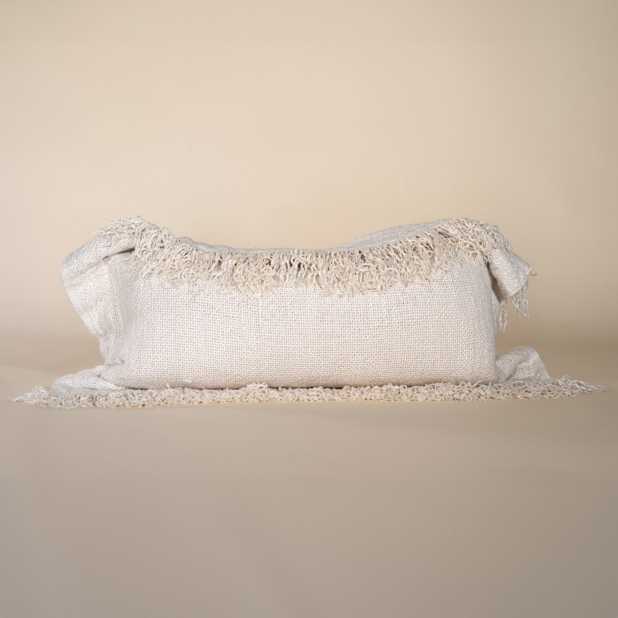 A natural-toned lumbar cushion with a woven texture, featuring delicate fringed edges, set against a neutral backdrop