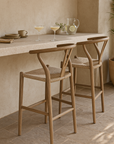 Haani Teak Bar Chair styled in a Mediterranean-inspired outdoor setting, featuring a curved teak frame and woven seat at a rendered stone counter with natural tableware and drinks.