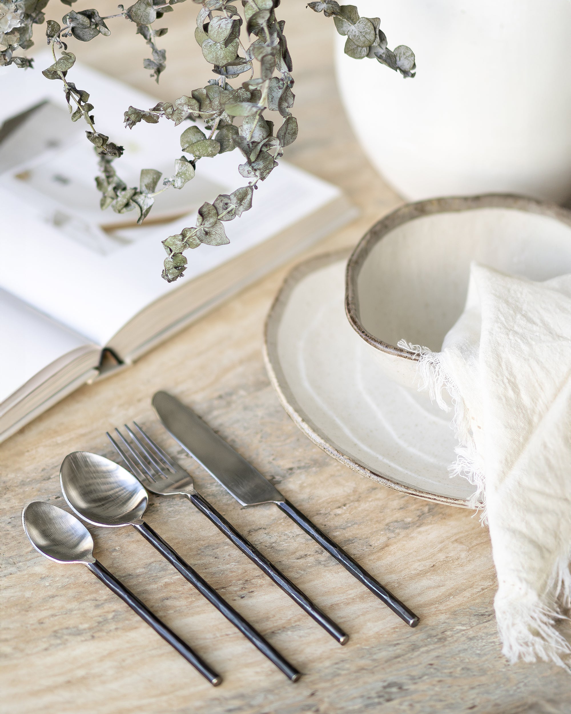 A minimalist table setting featuring hand-forged stainless steel cutlery with sleek black handles, paired with rustic ceramic dishware on a textured stone table, evoking a refined yet organic dining experience