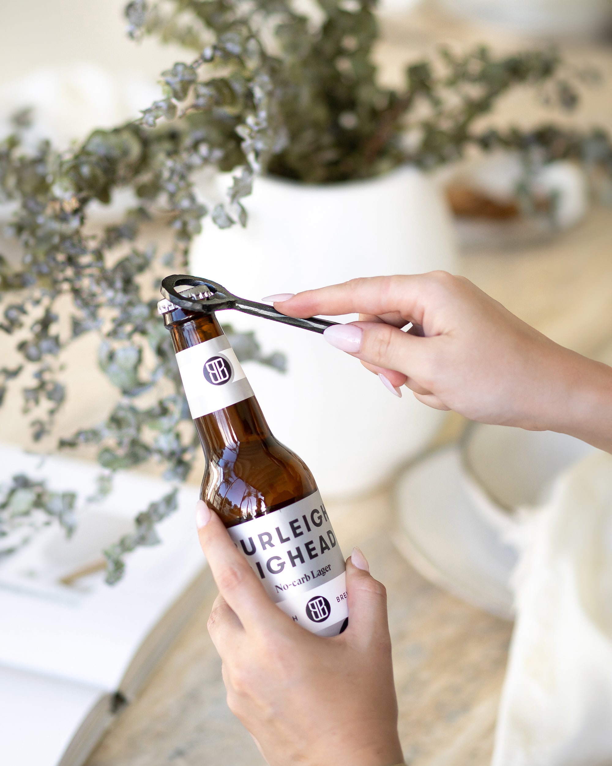 A hand-forged black iron bottle opener in use, effortlessly opening a Burleigh Bighead beer bottle against a stylish neutral-toned setting with a white ceramic vase and dried foliage