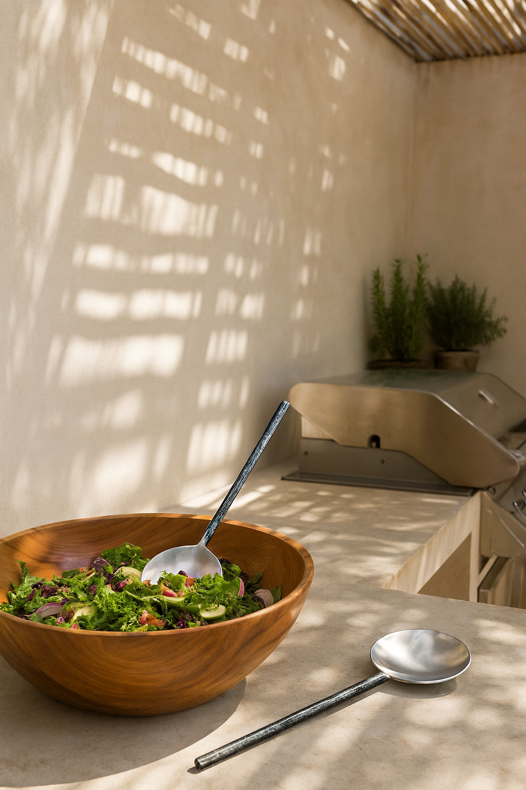 Handcrafted Artisan Salad Servers in forged stainless steel styled with a timber salad bowl in an outdoor kitchen setting with natural light and textured walls