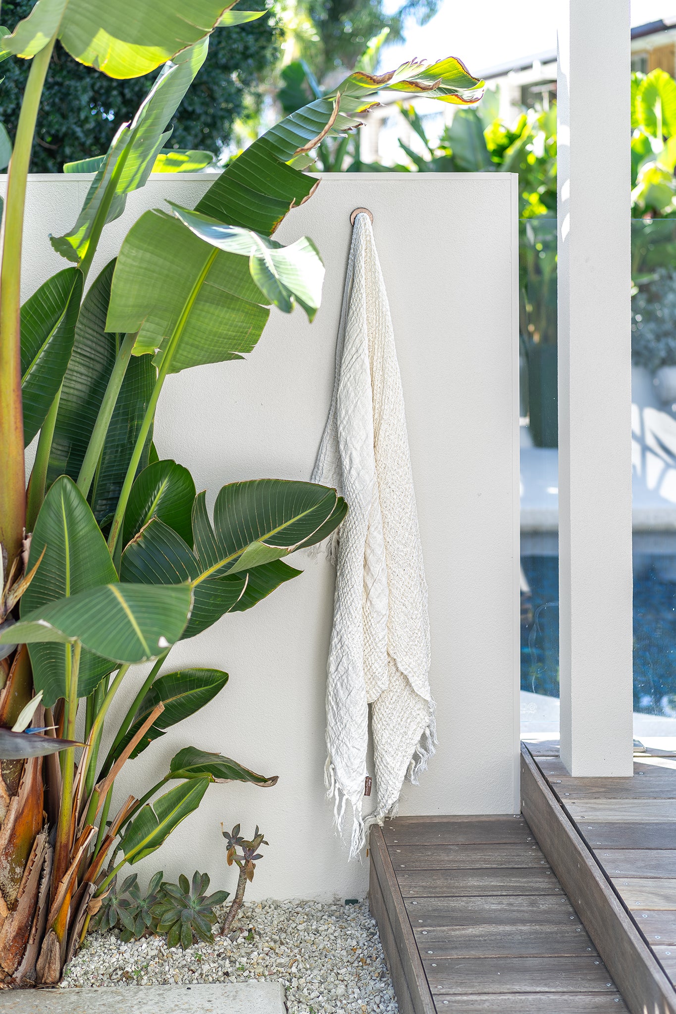Ivory handloom linen bath towel hanging on an outdoor wall beside tropical foliage and timber decking, capturing a relaxed resort-style atmosphere by the pool.