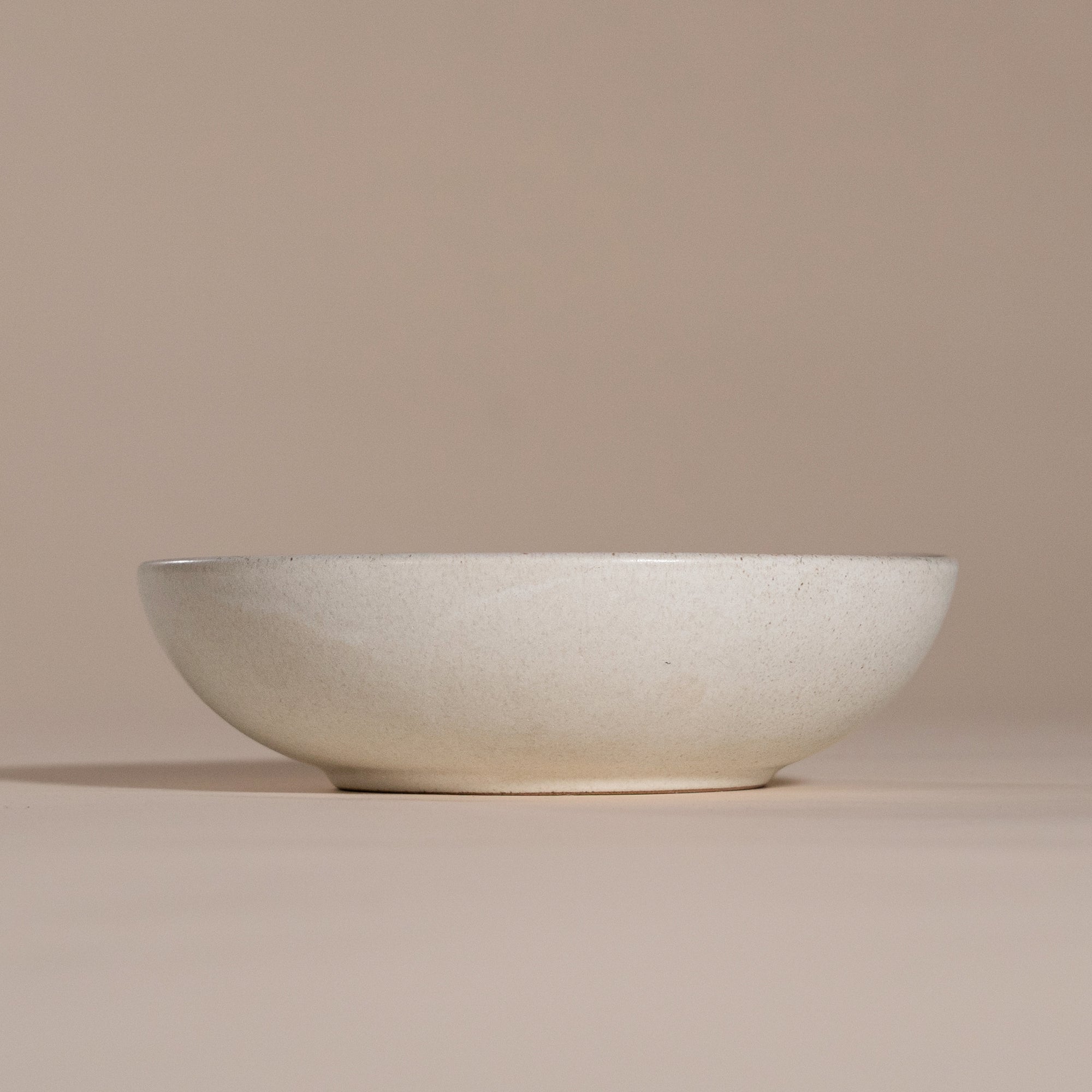 A handmade ceramic bowl with a smooth, speckled neutral glaze, set against a soft beige background. The minimalist design and organic shape make it a versatile piece for modern dining and home decor