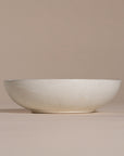 A handmade ceramic bowl with a smooth, speckled neutral glaze, set against a soft beige background. The minimalist design and organic shape make it a versatile piece for modern dining and home decor