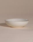 A handmade ceramic bowl with a smooth, speckled neutral glaze, set against a soft beige background. The minimalist design and organic shape make it a versatile piece for modern dining and home decor