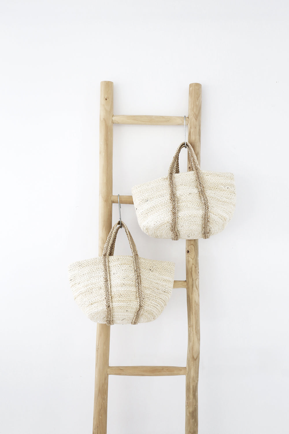 Two handwoven natural fibre market bags with braided handles hanging on a rustic wooden ladder against a white wall. A minimalist coastal-inspired storage solution with organic textures