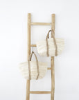 Two handwoven natural fibre market bags with braided handles hanging on a rustic wooden ladder against a white wall. A minimalist coastal-inspired storage solution with organic textures