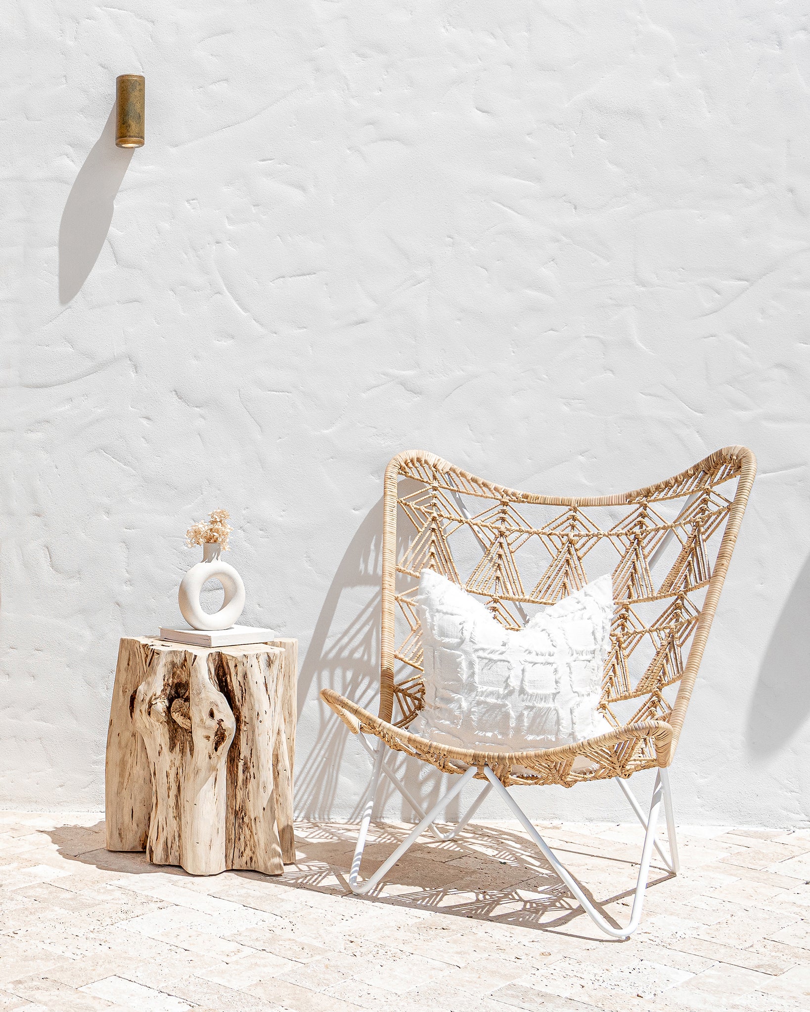 A serene Mediterranean-inspired outdoor space featuring a rattan butterfly chair with a white frame, a natural tree stump side table, and a textured cushion. The open timber doors reveal a breezy indoor lounge with white drapes, a sculptural olive tree in a white planter, and organic wood furniture, bathed in natural sunlight