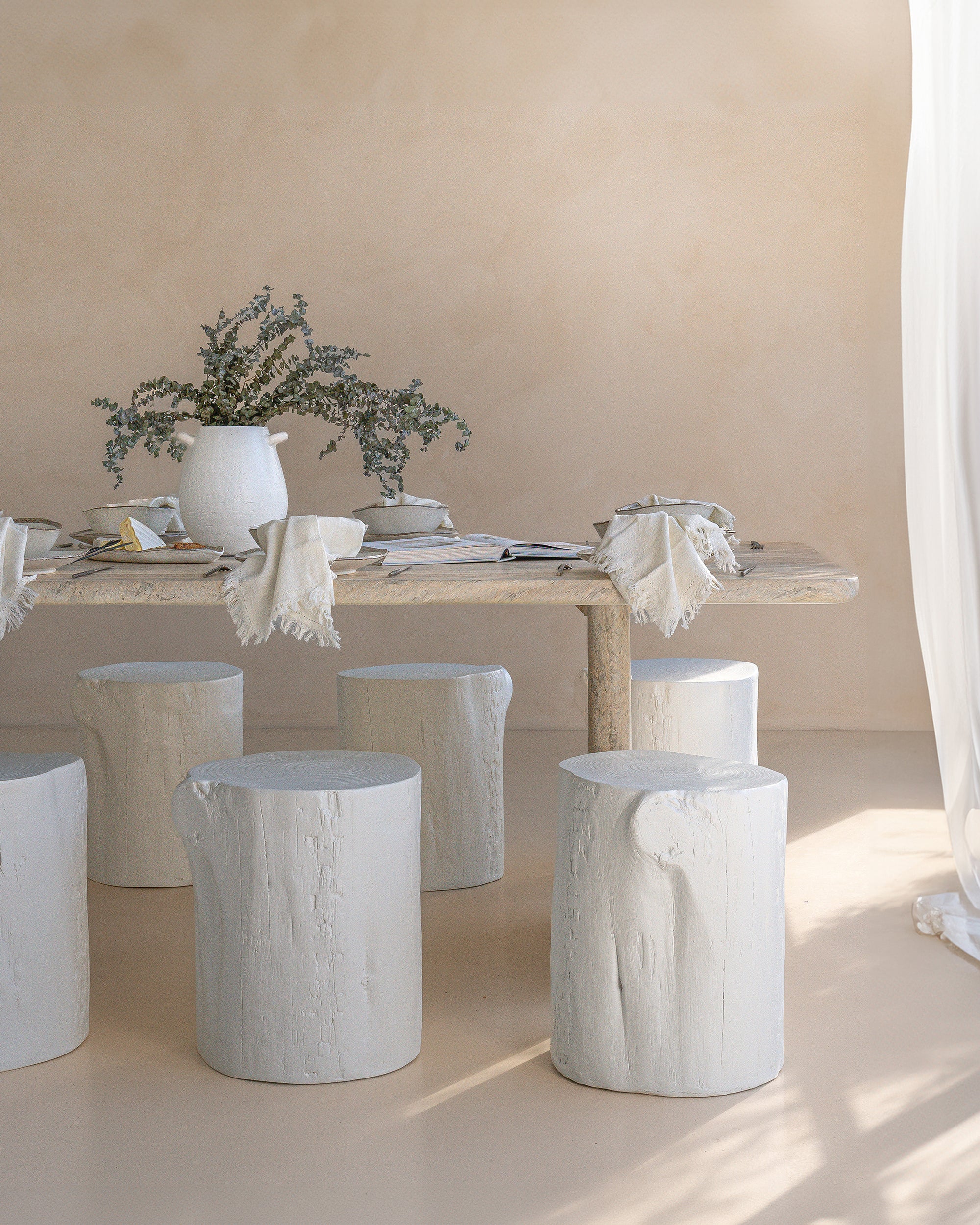 Hyrdo-dipped dining table styled with white timber stump stools, neutral linen napkins, and ceramic tableware. A soft, sunlit setting with organic textures and natural elements, creating a relaxed, earthy ambiance