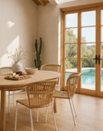 Indoor-outdoor dining chairs with natural rope seats and white metal legs styled around an oak dining table in a sunlit room with a poolside view