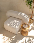 A sculptural ivory bouclé armchair with a rounded silhouette, styled with a handcrafted wooden side table. Sunlight filters through greenery, casting soft shadows in this serene and minimalist setting.