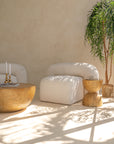 A sculptural ivory bouclé armchair with a rounded silhouette, styled with a handcrafted wooden side table. Sunlight filters through greenery, casting soft shadows in this serene and minimalist setting