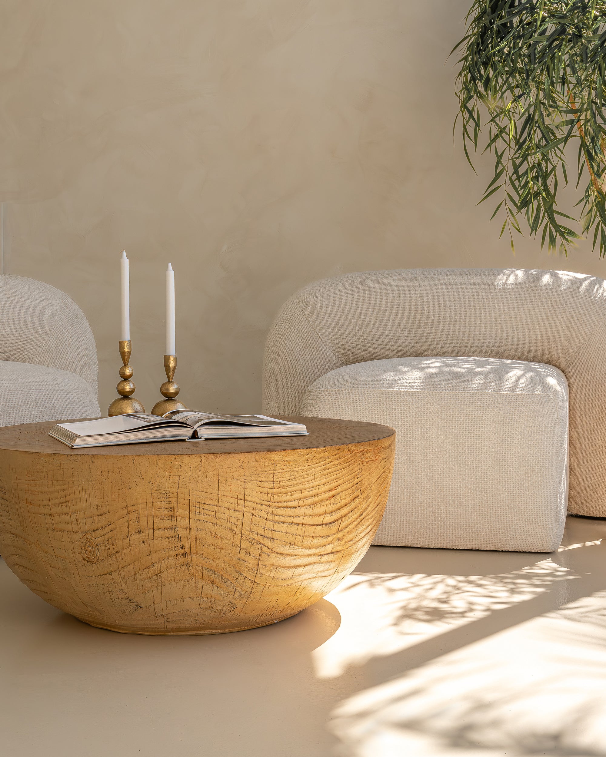 A sculptural ivory bouclé armchair with a rounded silhouette, styled with a handcrafted wooden side table. Sunlight filters through greenery, casting soft shadows in this serene and minimalist setting
