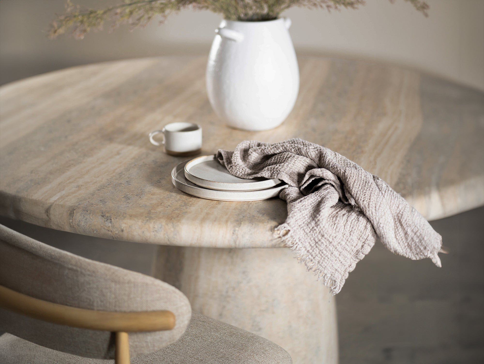 A close-up of a travertine dining table paired with a seat chair. The natural stone surface is complemented mug of water, and soft decorative foliage, creating a warm, organic aesthetic
