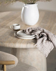 A close-up of a travertine dining table paired with a seat chair. The natural stone surface is complemented mug of water, and soft decorative foliage, creating a warm, organic aesthetic