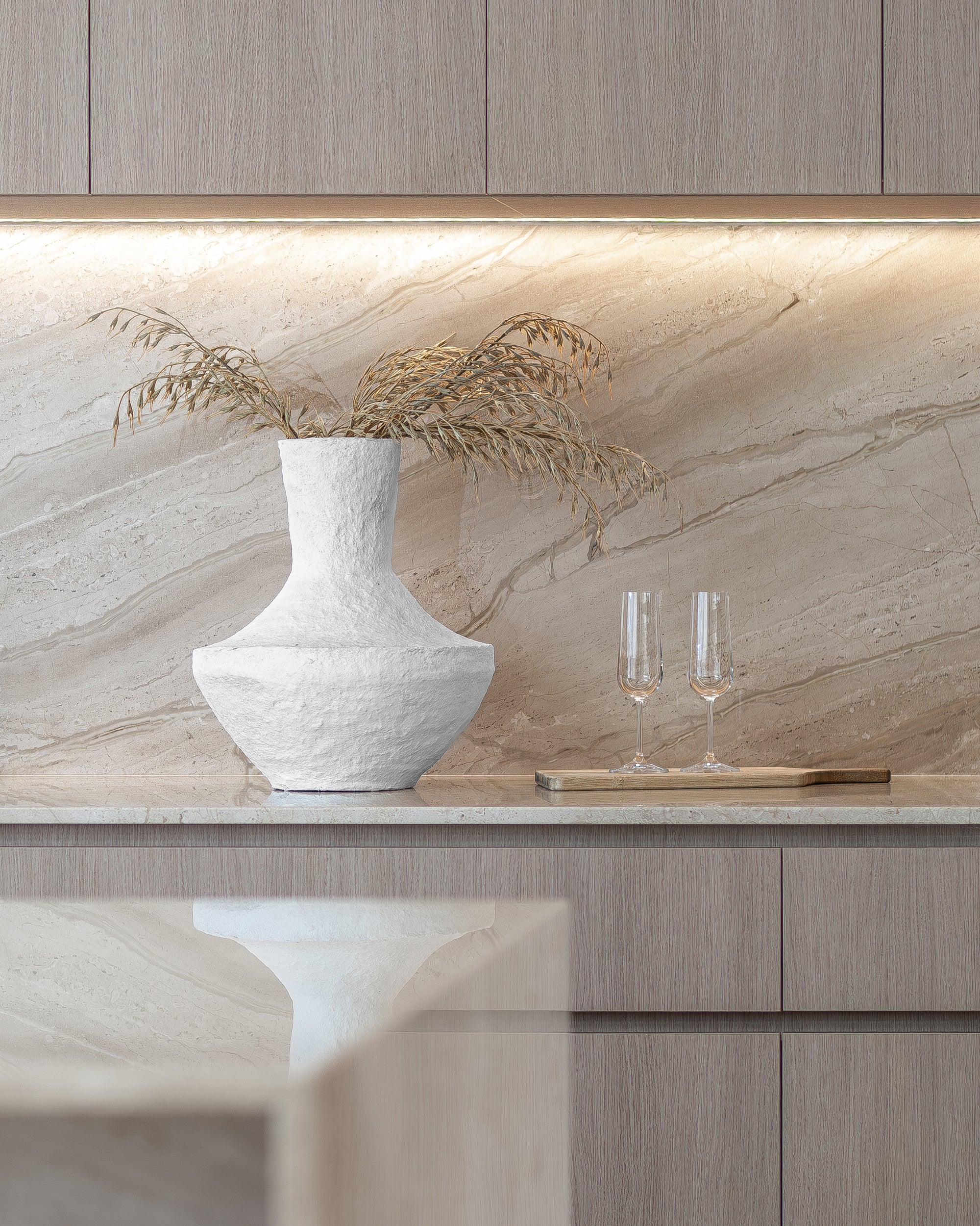 Modern kitchen styling featuring a sculptural white textured vase with dried foliage and two champagne flutes on a marble benchtop with soft under-cabinet lighting.