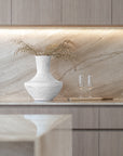 Modern kitchen styling featuring a sculptural white textured vase with dried foliage and two champagne flutes on a marble benchtop with soft under-cabinet lighting.