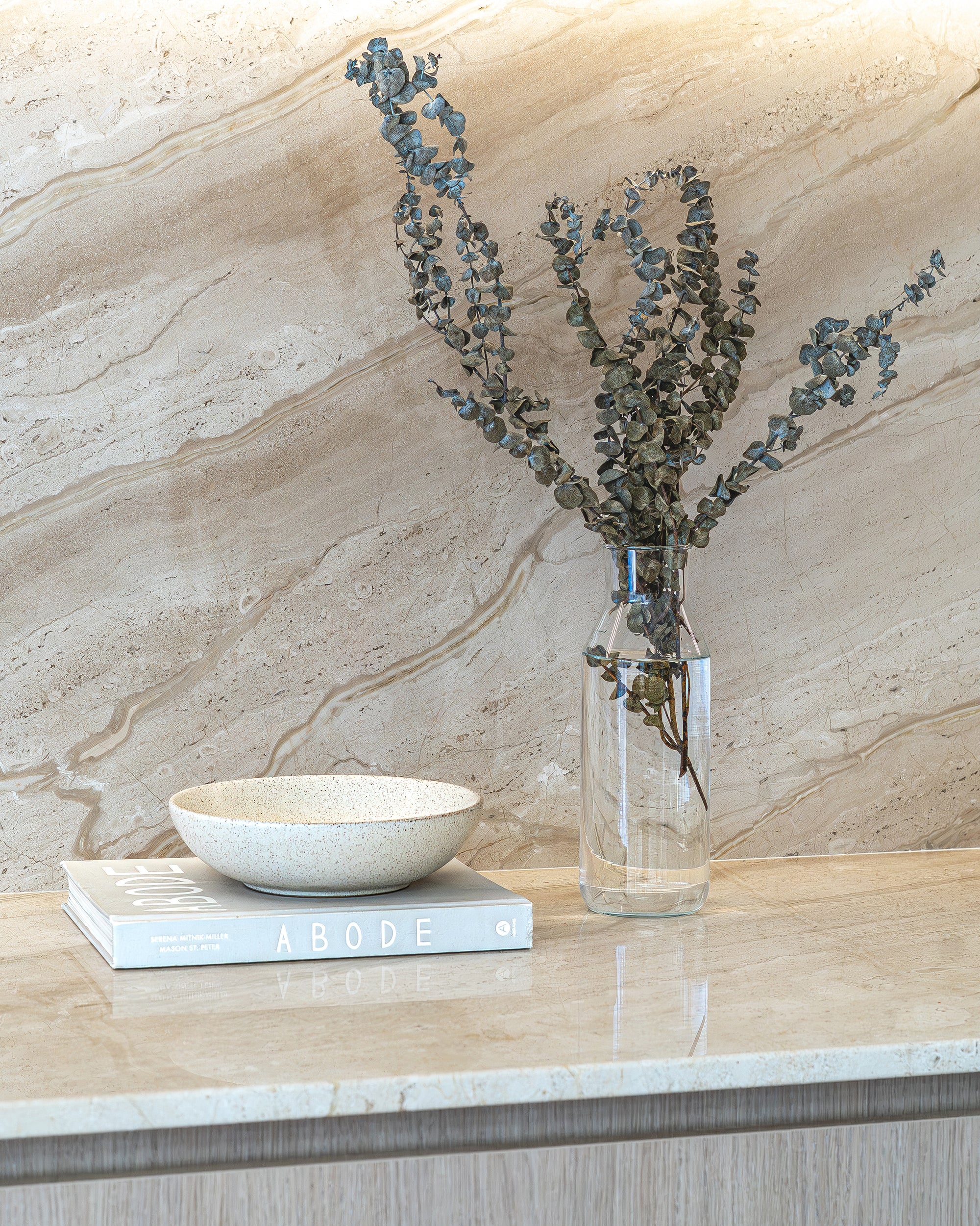 Neutral-toned marble benchtop styled with a clear glass vase of dried eucalyptus, a ceramic bowl, and the Abode book in a minimalist kitchen setting.