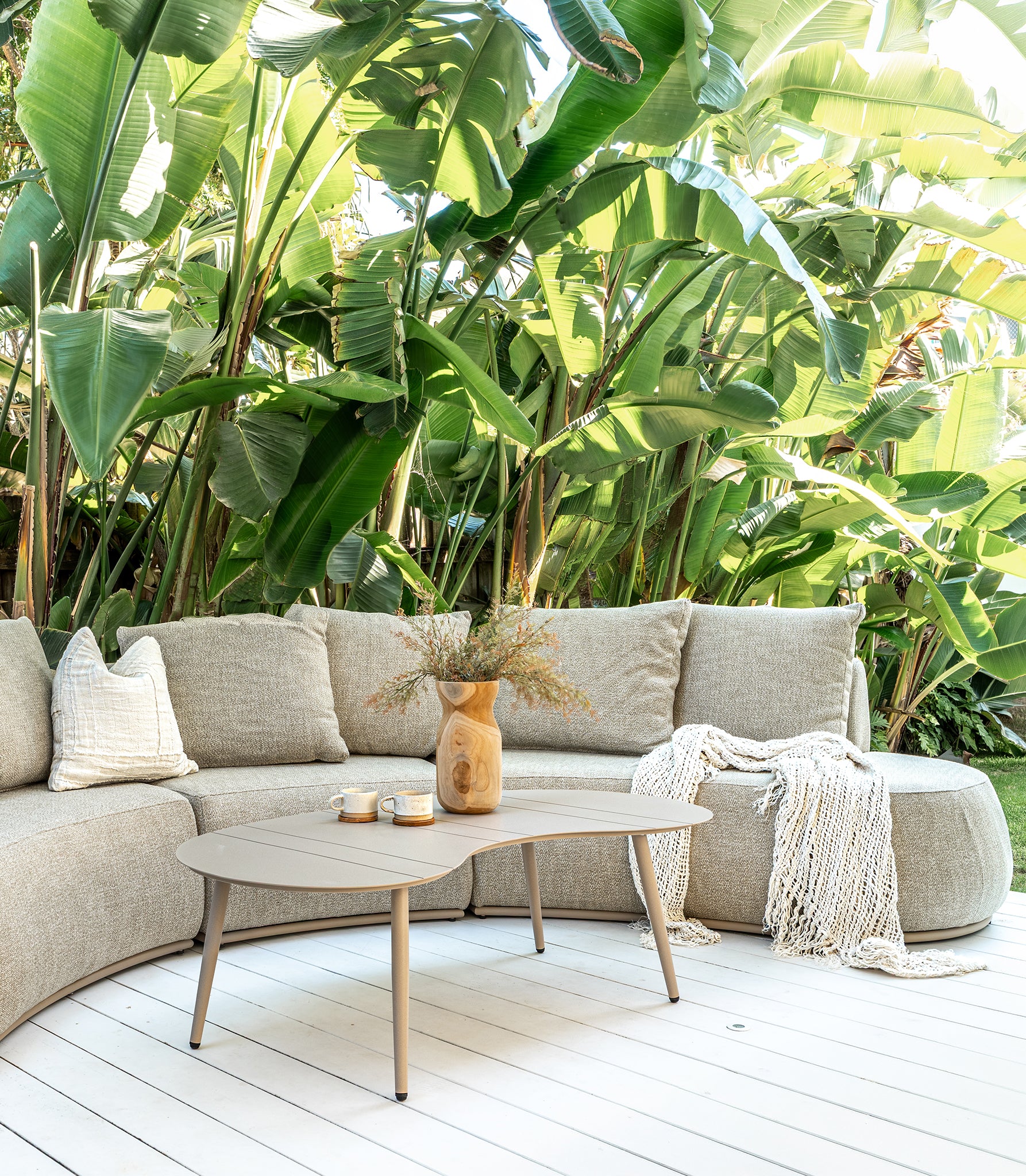 Laguna outdoor sofa in oat drift fabric with Solara coffee table on a sunlit deck surrounded by lush tropical greenery creating a relaxed resort style setting