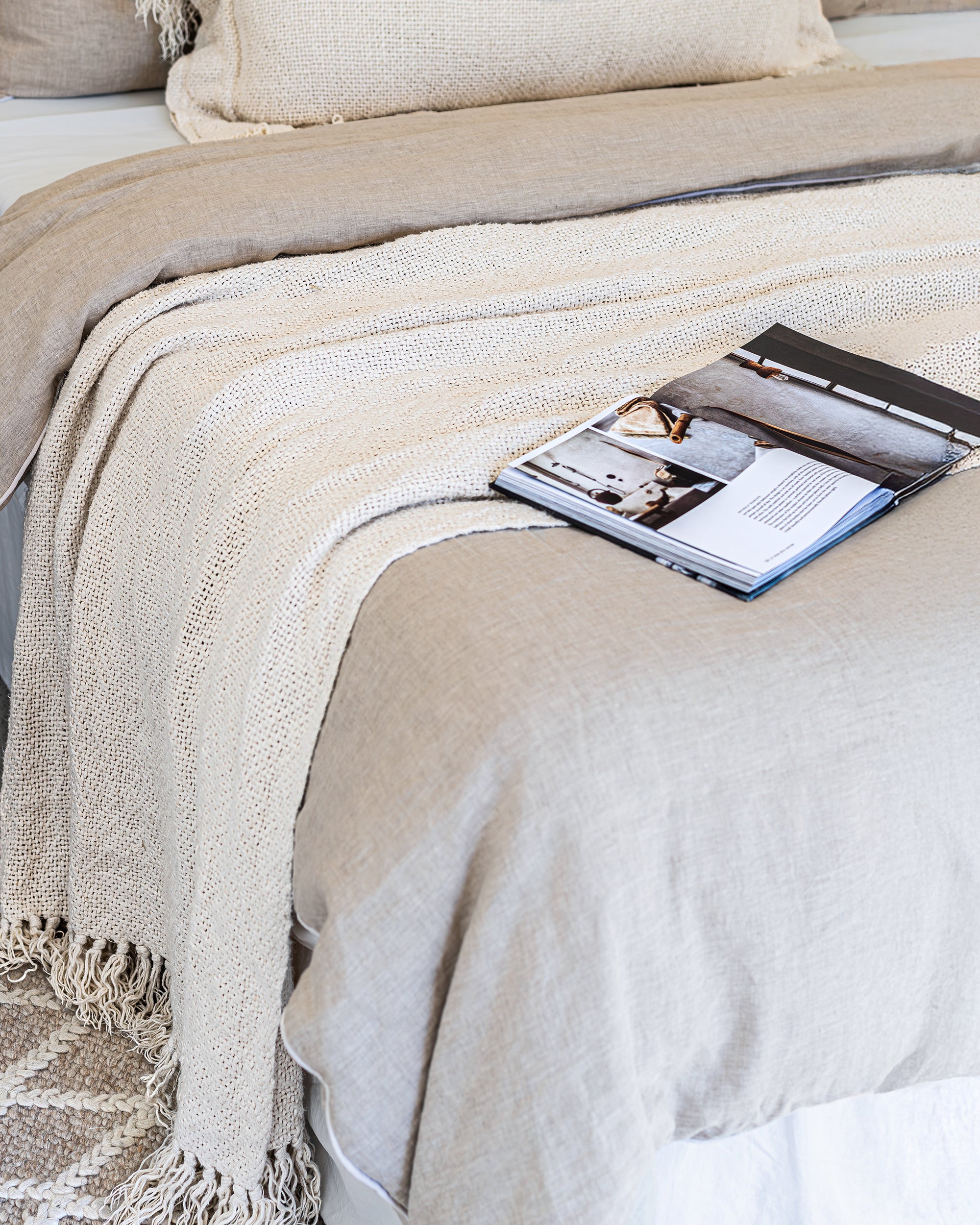 Detail of layered linen bedding and a chunky knit throw in soft neutral tones, styled with an open magazine on a textured wool floor rug