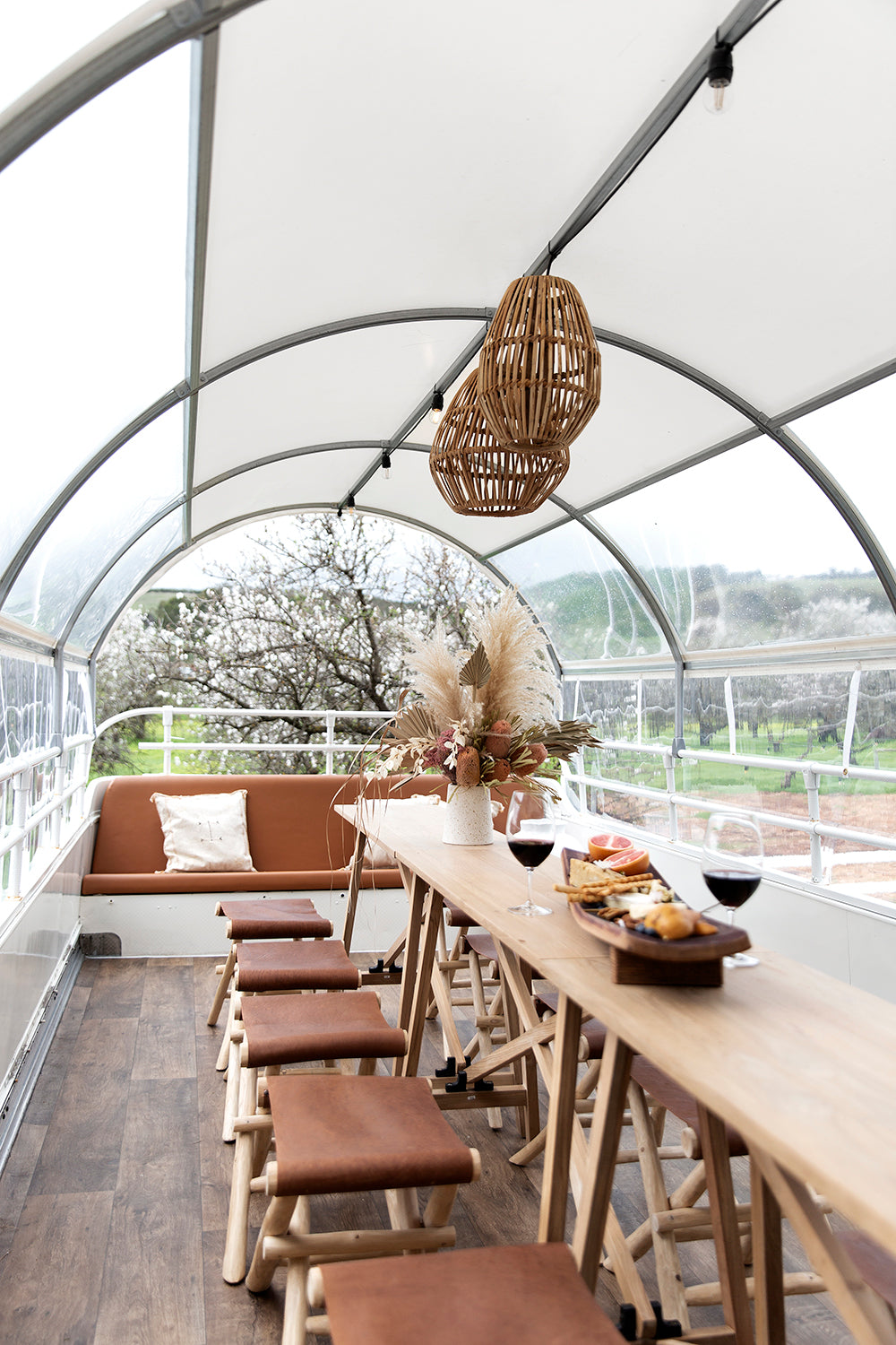 Outdoor entertaining space with natural teak wood console table, leather seat stools, rattan pendant lights, and floral centrepiece under a curved canopy