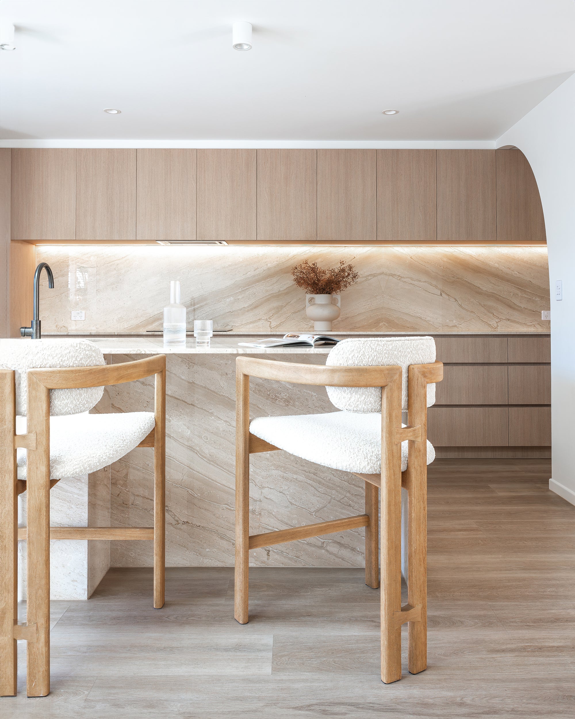 Pair of light oribberwood and boucle counter stools at a marble island bench in a contemporary kitchen with timber cabinetry and natural stone splashback.