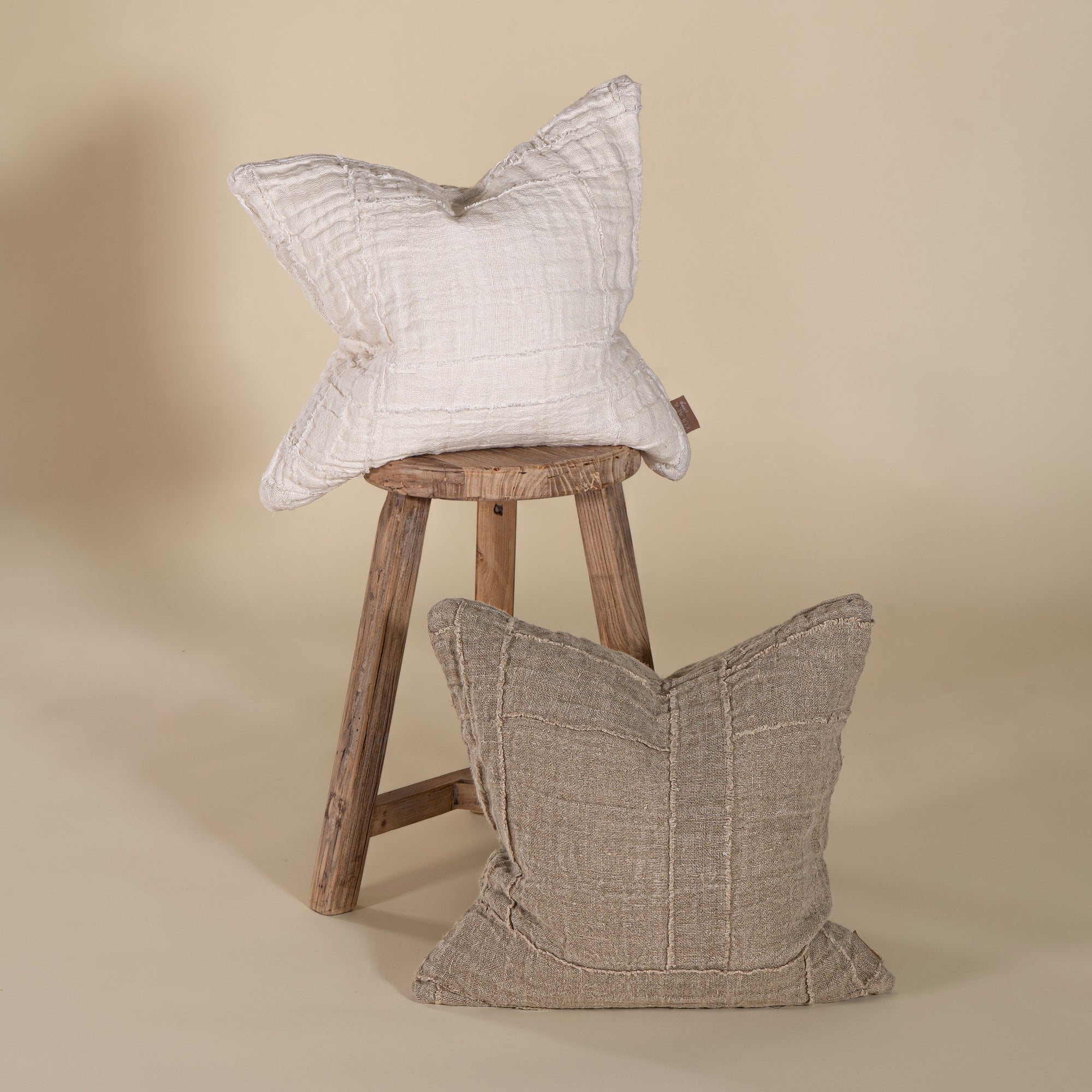 Set of handcrafted linen cushions in off-white and natural tones, featuring a textured patchwork design, styled on a rustic wooden stool