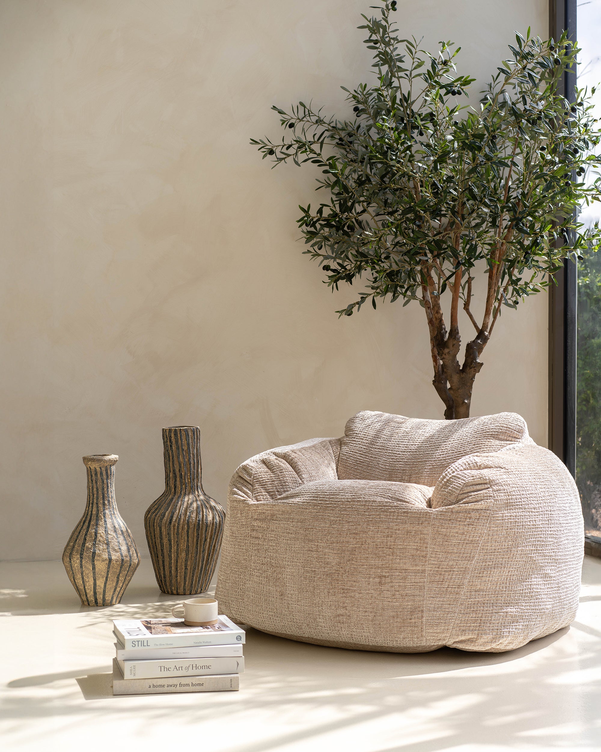 The Kulala Living luxe boucle bean bag in a serene, sunlit setting, styled with earthy-toned decor and an olive tree. Featuring a soft natural texture, this oversized bean bag blends modern comfort with relaxed elegance, perfect for a tranquil living space