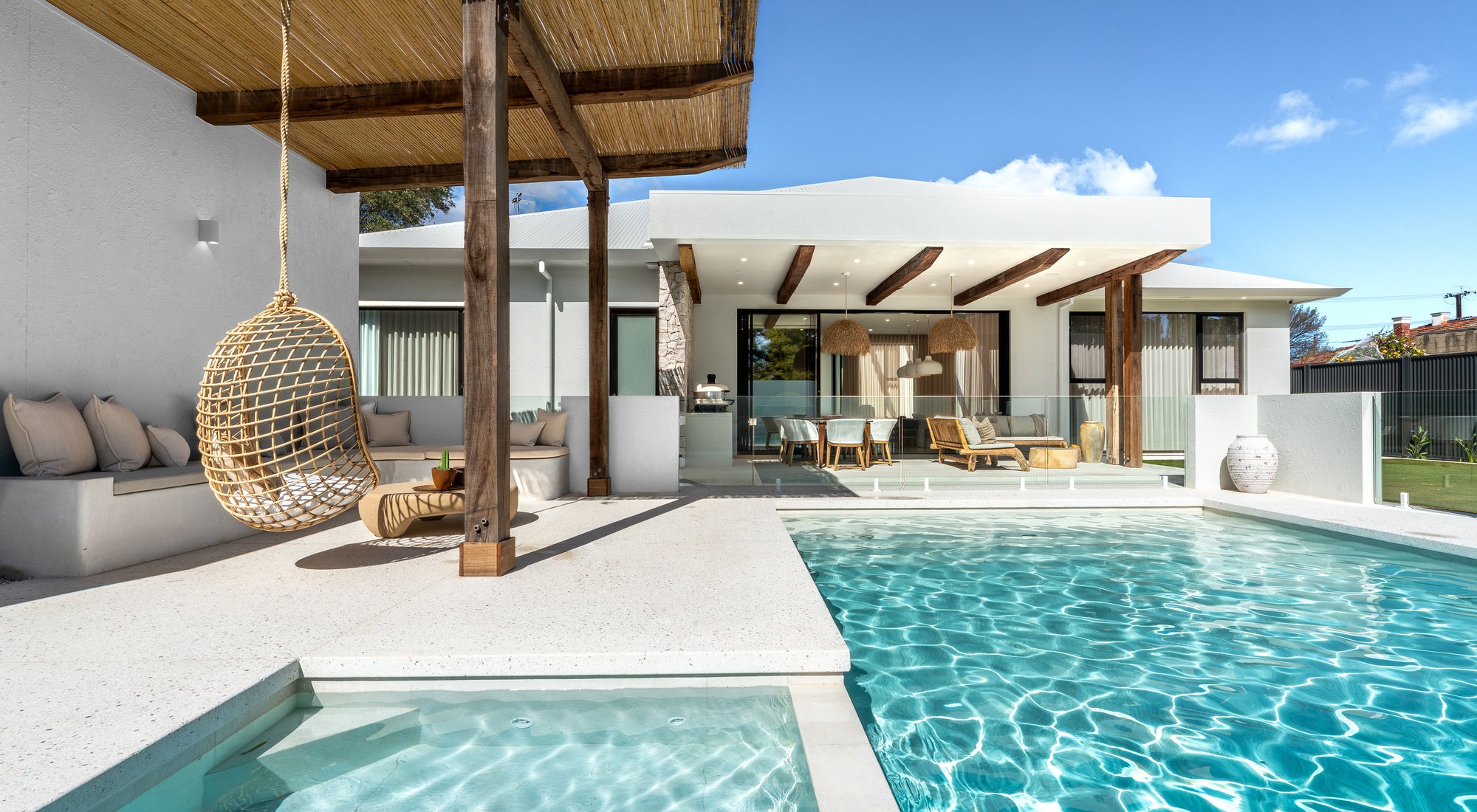 Luxury outdoor poolside lounge with rattan hanging chair, timber pergola, cushioned built-in seating and dining space overlooking the pool.