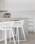 A bright modern kitchen featuring Kulala Living Makena Bar Chairs in crisp white, offering a sculptural rattan backrest and plush seating. Styled with neutral tones, natural wood cabinetry, and fresh greenery for an inviting, coastal-inspired space