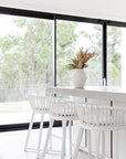 A bright modern kitchen featuring Kulala Living Makena Bar Chairs in crisp white, offering a sculptural rattan backrest and plush seating. Styled with neutral tones, natural wood cabinetry, and fresh greenery for an inviting, coastal-inspired space
