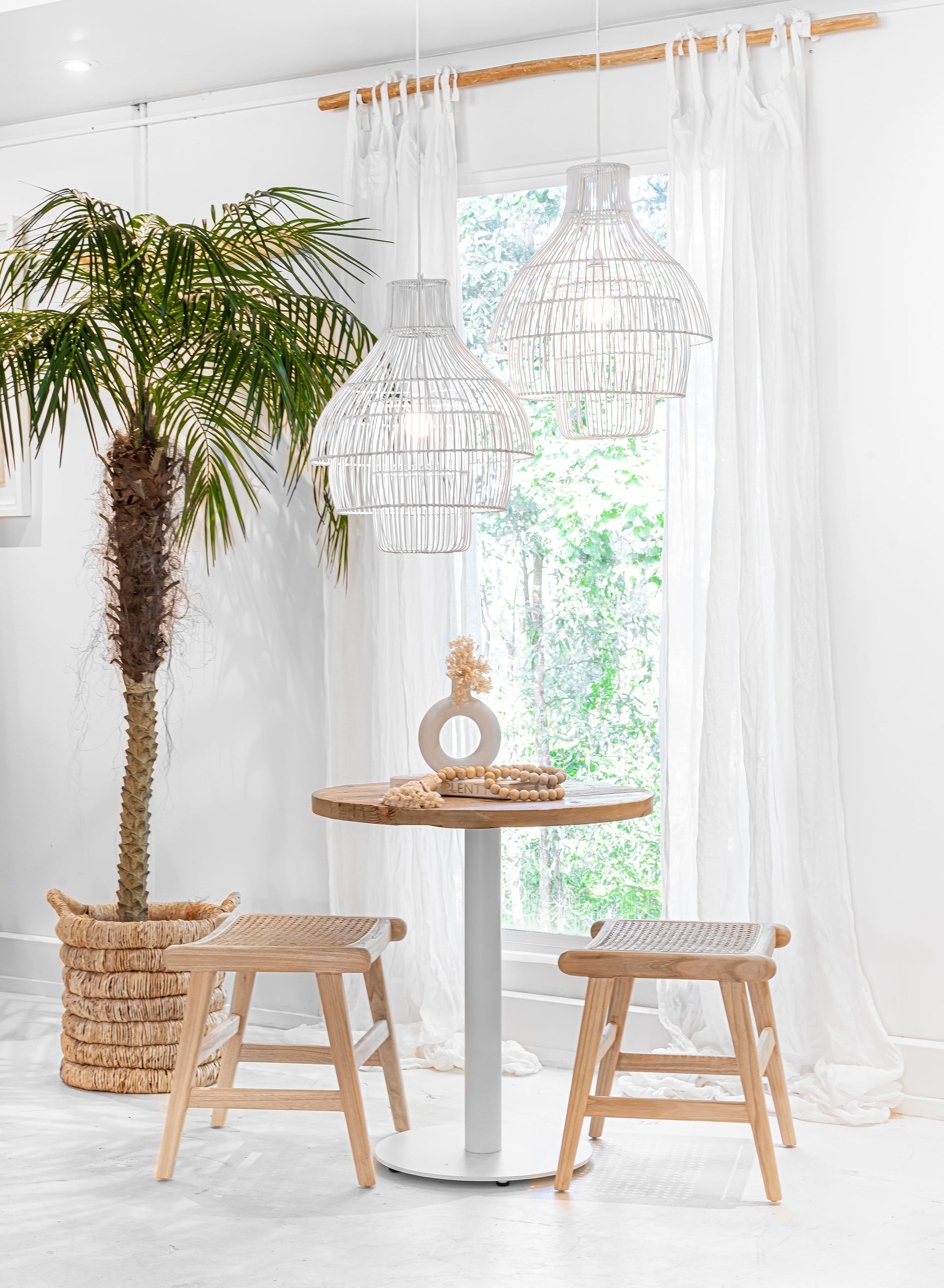 Mara basket styled as a woven planter beside a round timber table with stools in a bright coastal interior with white curtains and rattan pendant lights.