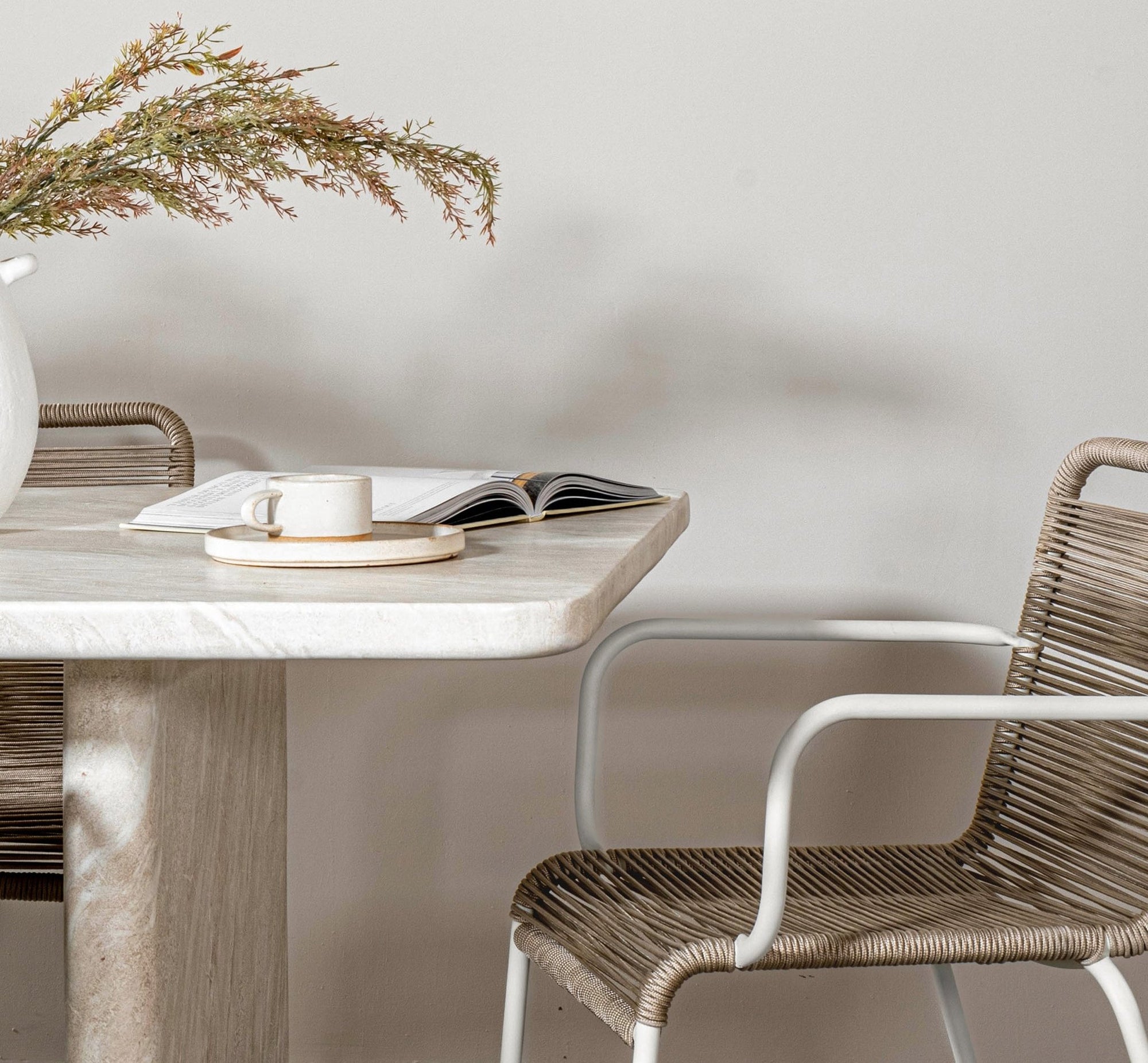 A serene dining setting featuring a marble dining table paired with a woven rope dining chair with a white metal frame. A handmade ceramic espresso cup sits on a matching saucer, complemented by an open book and a white ceramic vase with natural foliage. A perfect blend of organic textures and contemporary elegance
