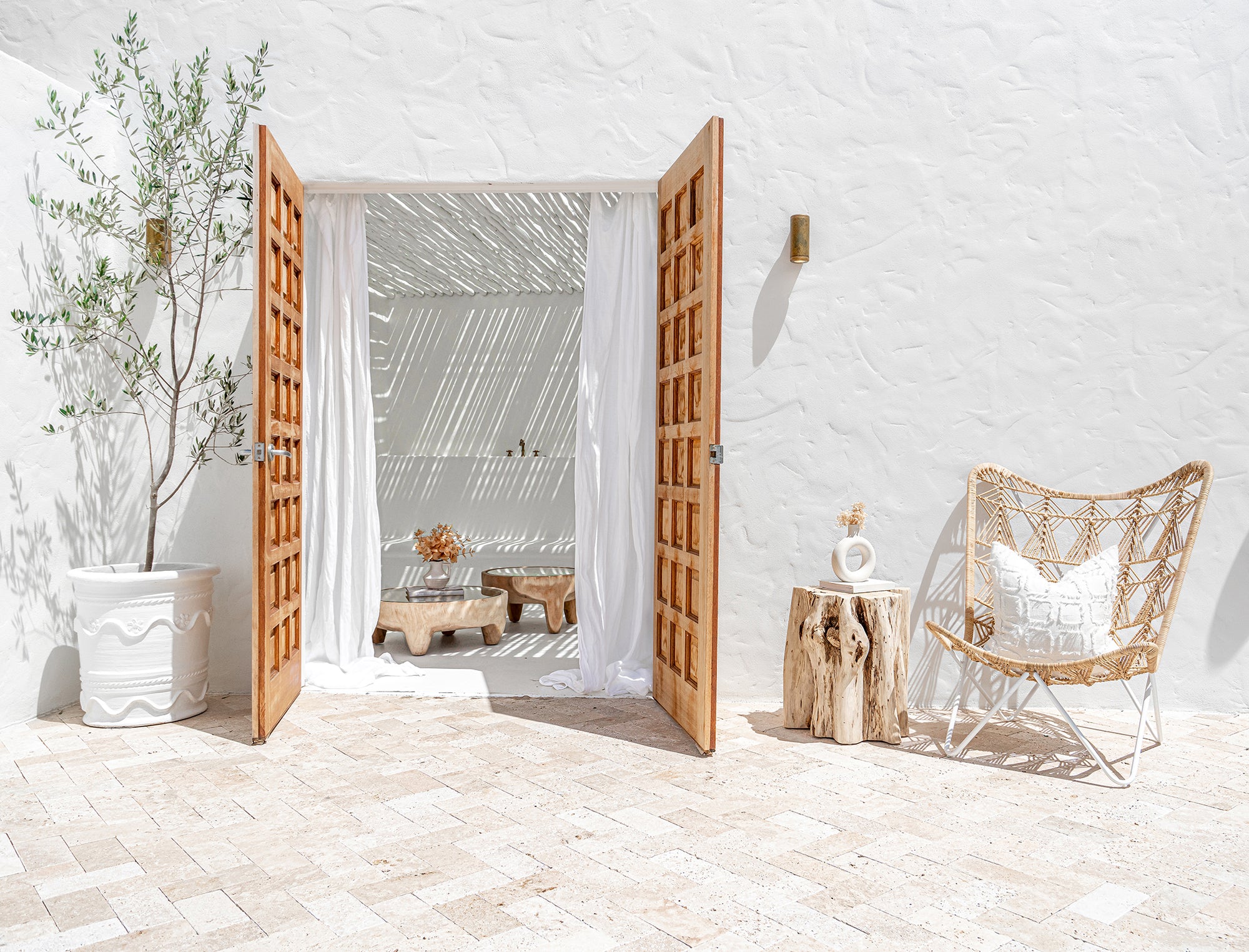 A serene Mediterranean-inspired outdoor space featuring a rattan butterfly chair with a white frame, a natural tree stump side table, and a textured cushion. The open timber doors reveal a breezy indoor lounge with white drapes, a sculptural olive tree in a white planter, and organic wood furniture, bathed in natural sunlight