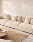 Melonee modular sofa in soft beige upholstery styled in a warm neutral living room with plaster walls, woven rug and round timber coffee table.