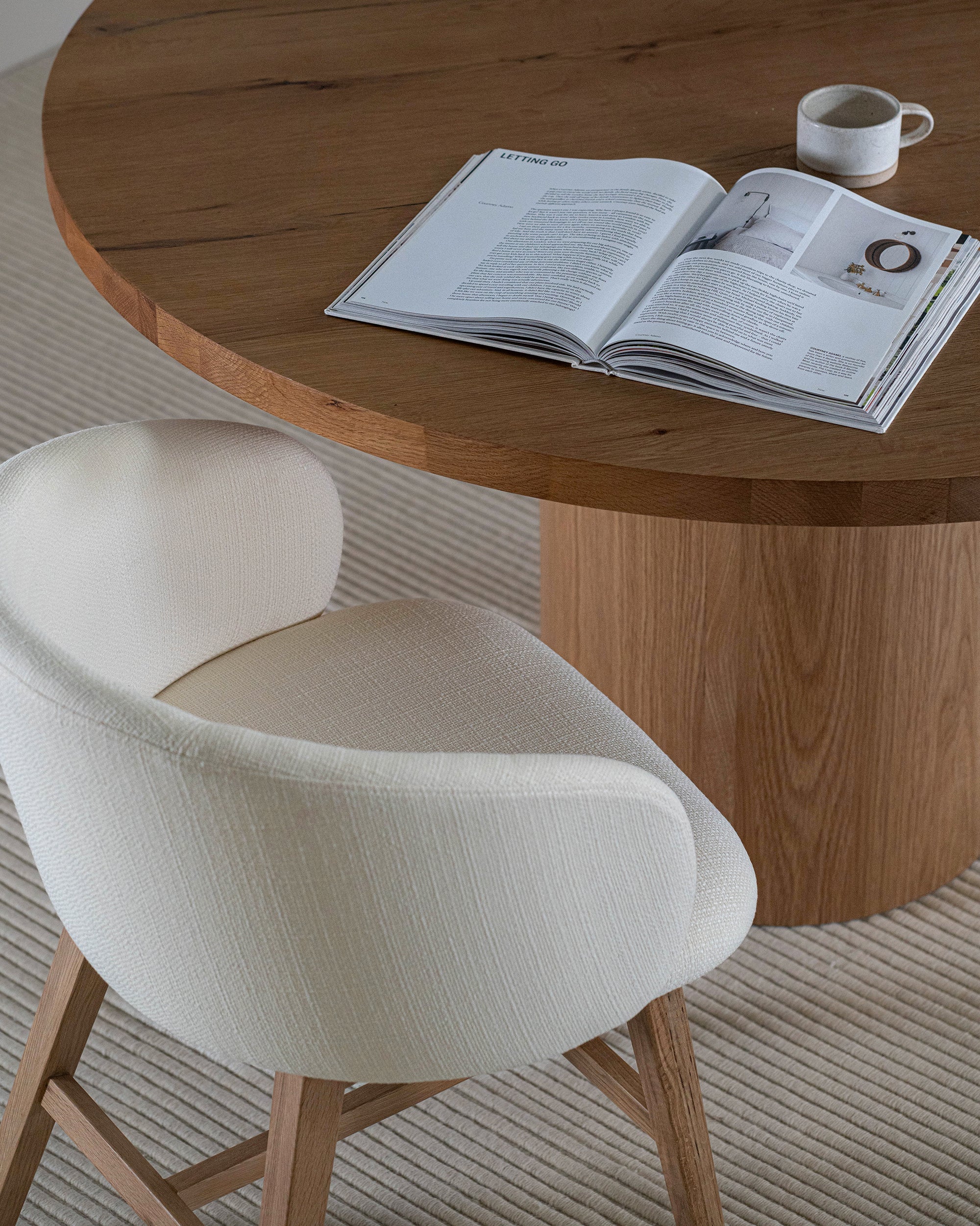 A contemporary upholstered dining chair with a curved backrest and oak timber legs, styled with a round wooden dining table in a neutral-toned interior