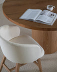 A contemporary upholstered dining chair with a curved backrest and oak timber legs, styled with a round wooden dining table in a neutral-toned interior