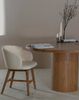 A contemporary upholstered dining chair with a curved backrest and oak timber legs, styled with a round wooden dining table in a neutral-toned interior