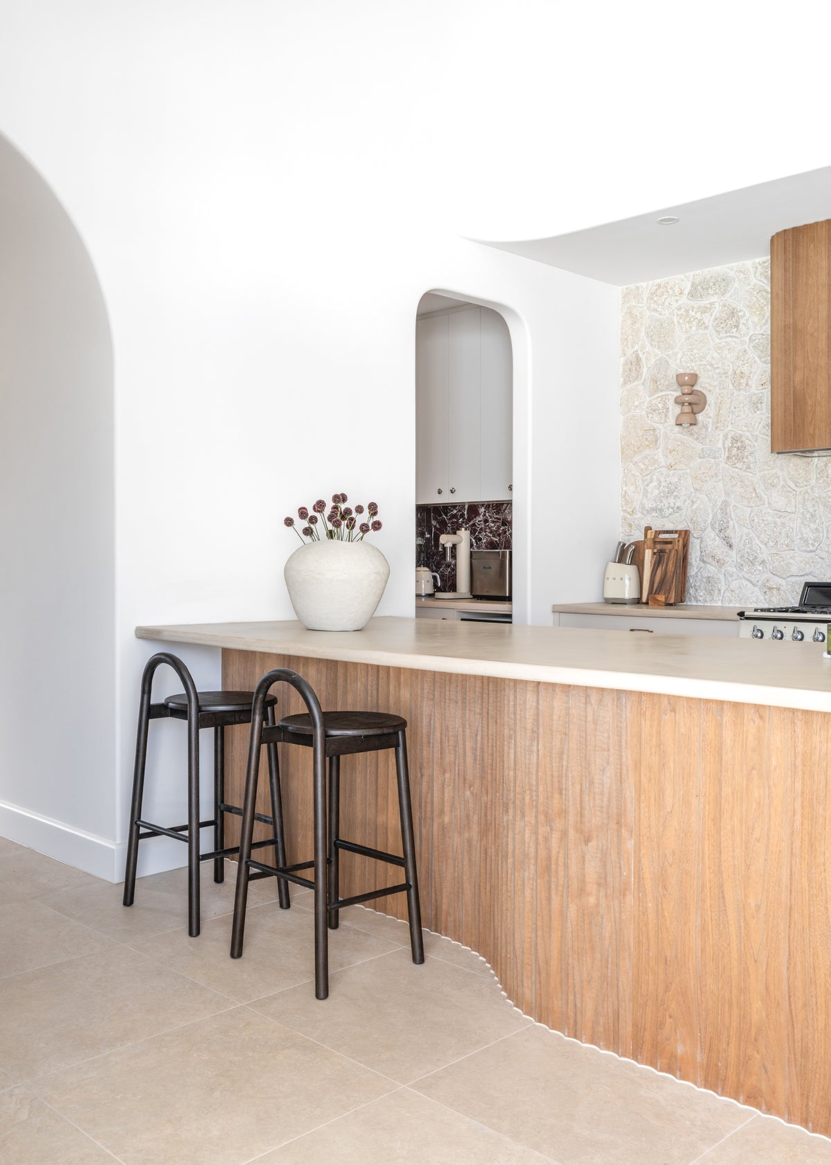 Two KULALA Living Milo Timber Bar Chairs in Espresso styled at a curved timber kitchen island with a natural stone benchtop, textured stone feature wall and warm coastal interiors.