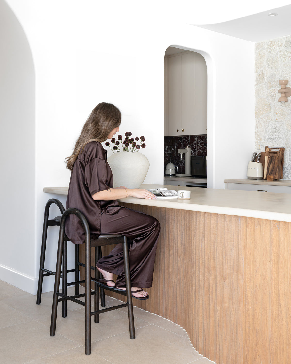KULALA Living Milo Timber Bar Chairs in Espresso and handcrafted ceramic mug in Sesame Glaze, styled in a light filled coastal kitchen with a curved timber island and natural stone benchtop.