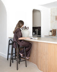 KULALA Living Milo Timber Bar Chairs in Espresso and handcrafted ceramic mug in Sesame Glaze, styled in a light filled coastal kitchen with a curved timber island and natural stone benchtop.