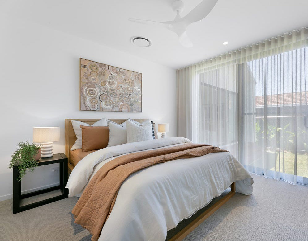 A serene modern bedroom featuring an oak bed frame dressed in soft white and earthy-toned bedding. Layered cushions and a quilted throw add warmth, complemented by sheer curtains and a statement artwork above the bed