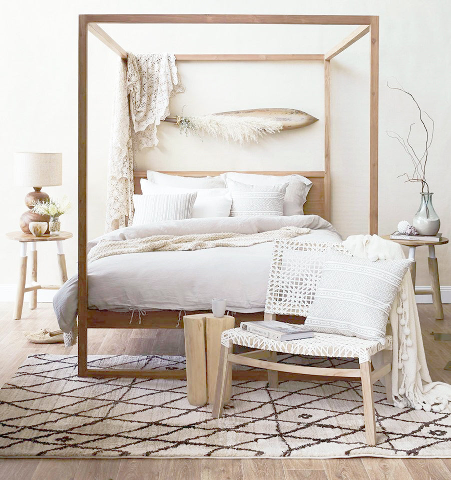 Light-filled modern boho bedroom with a wooden canopy bed, layered neutral bedding, woven accent chair with white rope weave, natural wood side tables, and organic decor accents