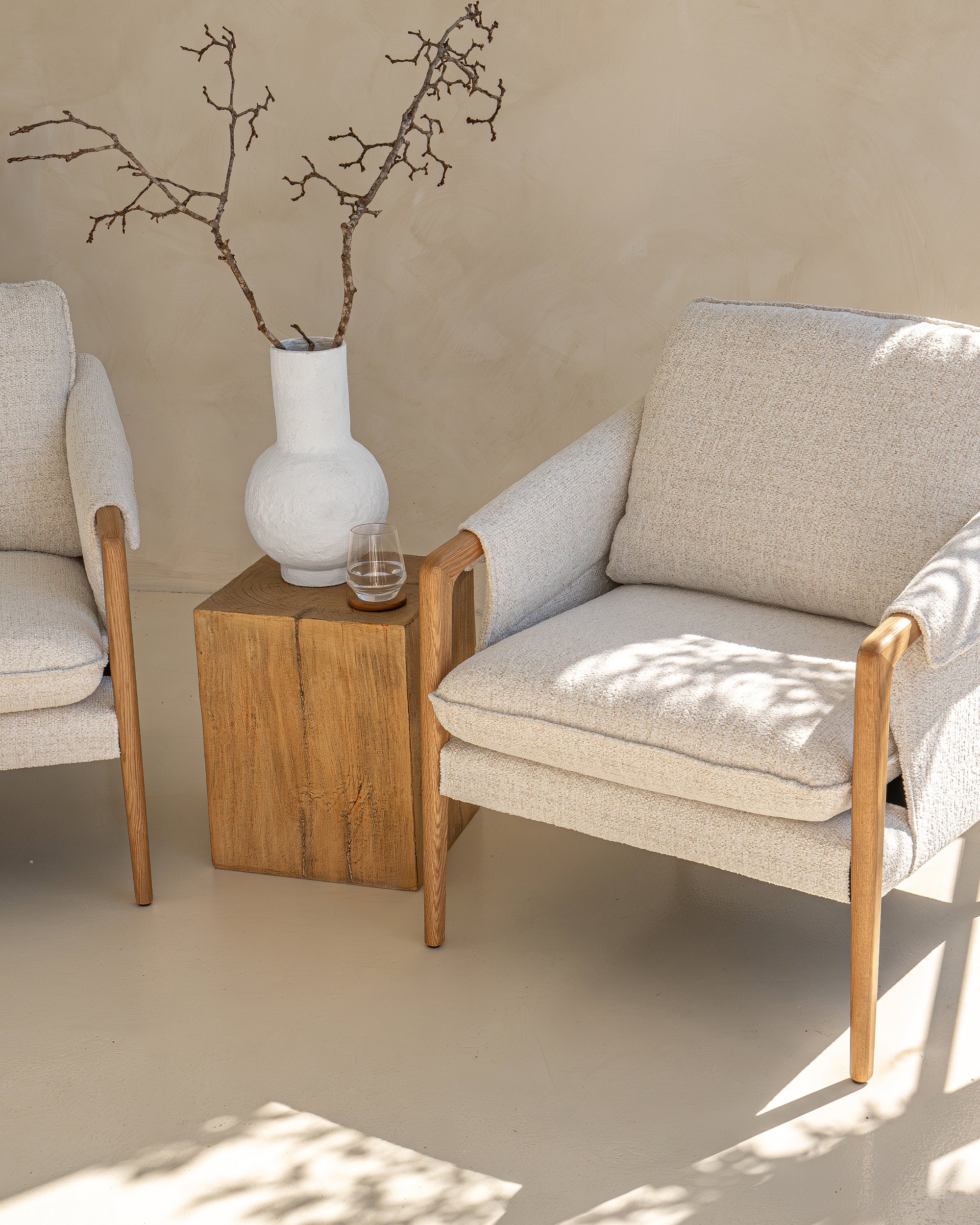 A serene living space featuring two modern boucle armchairs with natural wood frames, styled with a rustic magnesium oxide side table and a white textured vase. Bathed in soft natural light, this setting embodies effortless elegance and contemporary comfort.