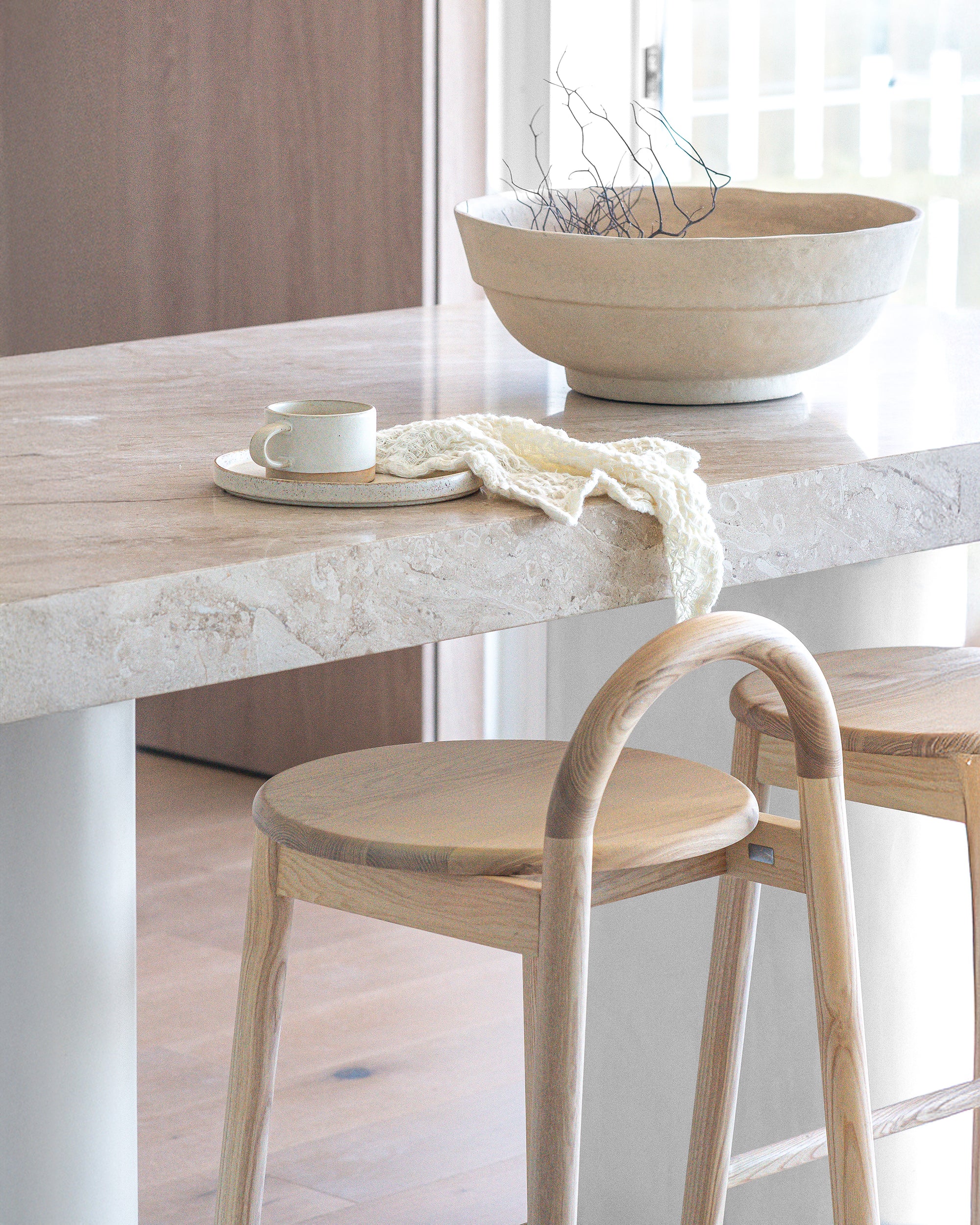 Natural timber stools at a travertine breakfast bar styled with a sculptural ceramic bowl, handmade cup and plate, and soft textured cloth in a light-filled KULALA Living kitchen setting