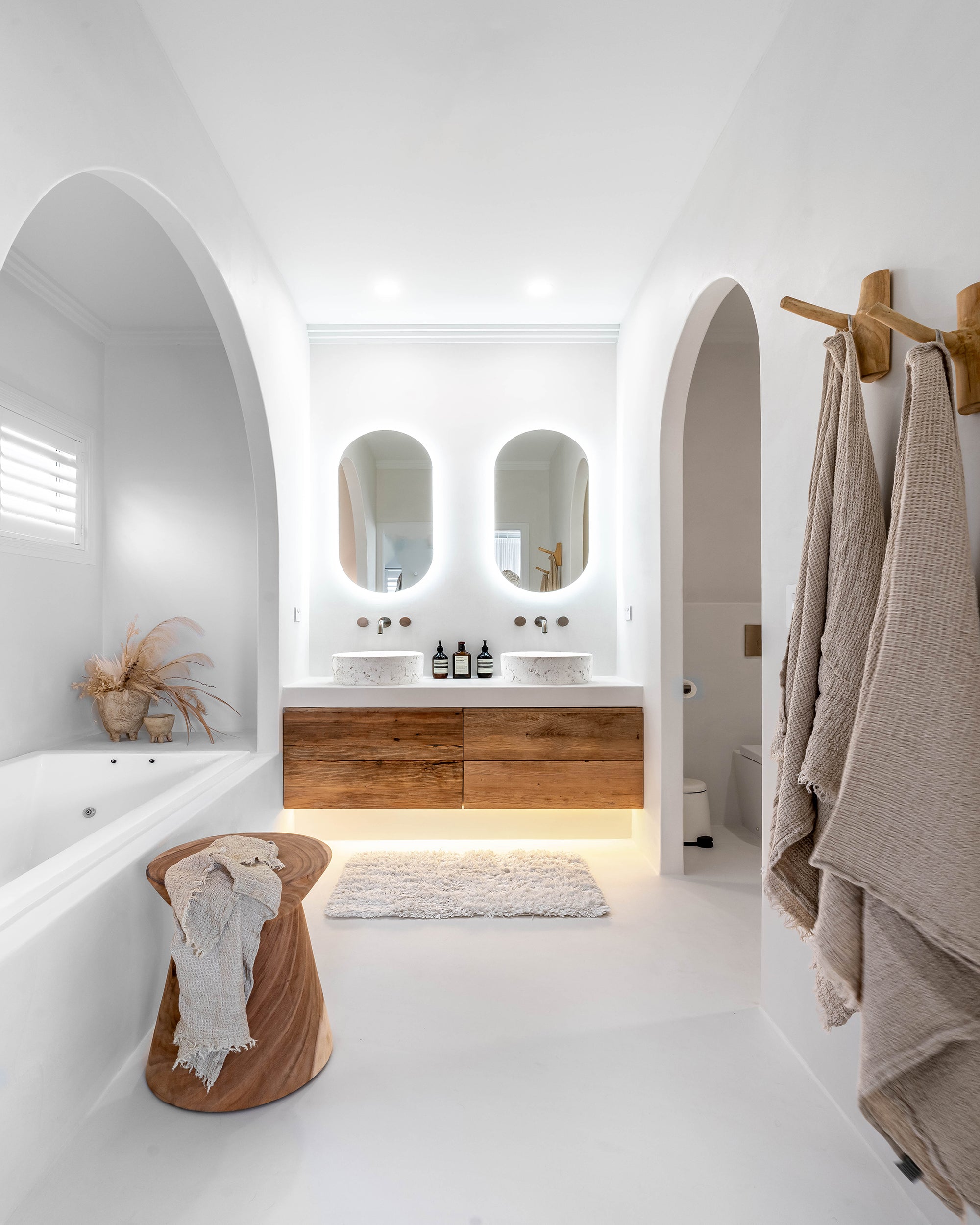 A serene modern coastal bathroom featuring natural wood elements, arched mirrors, and soft neutral tones. Includes a handcrafted wooden stool, wall hooks with linen towels, and backlit floating vanities for a minimalist, spa-like retreat